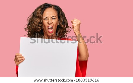 Middle age hispanic woman holding blank empty banner annoyed and frustrated shouting with anger, yelling crazy with anger and hand raised 