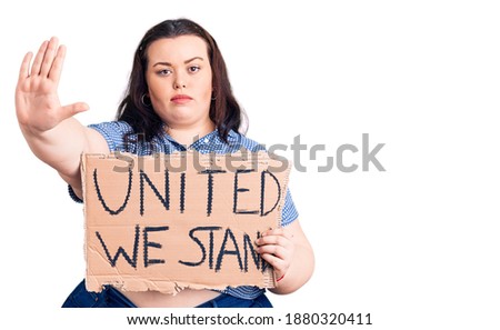 Young plus size woman holding united we stand banner with open hand doing stop sign with serious and confident expression, defense gesture 