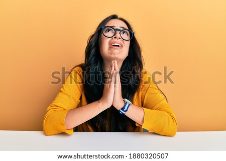 Beautiful brunette young woman wearing glasses and casual clothes sitting on the table begging and praying with hands together with hope expression on face very emotional and worried. begging. 