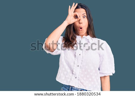 Young beautiful brunette woman wearing casual shirt doing ok gesture shocked with surprised face, eye looking through fingers. unbelieving expression. 