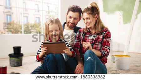 Portrait of happy little adorable Caucasian child girl sitting with young parents on floor after painting walls renovating home, typing and searching internet on tablet device, choosing room design.