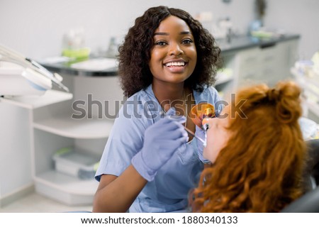 Smiling black woman dentist wearing blue uniform, providing tooth restoration and filling with curing polymerization UV lamp for her little patient, cute girl with red curly hair Royalty-Free Stock Photo #1880340133