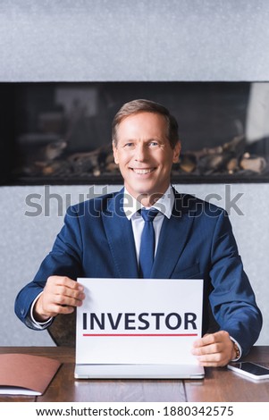 Smiling businessman looking at camera and holding sign with investor lettering while sitting at workplace in meeting room