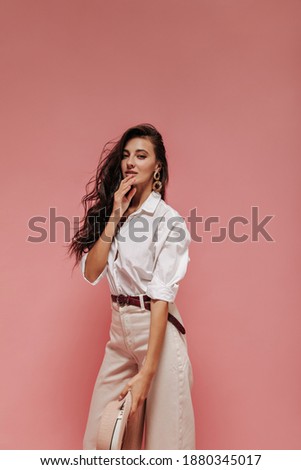 Charming girl with curly dark hair in gold earrings, white blouse, beige pants with brown belt looking into camera on pink backdrop..