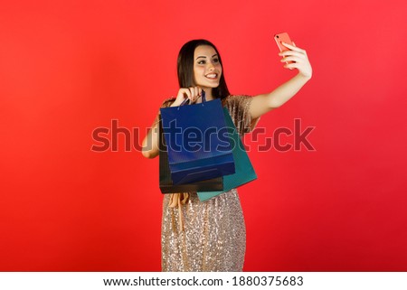 Beautiful happy girl makes selfie on her smartphone with shopping bags after shopping on red background. 