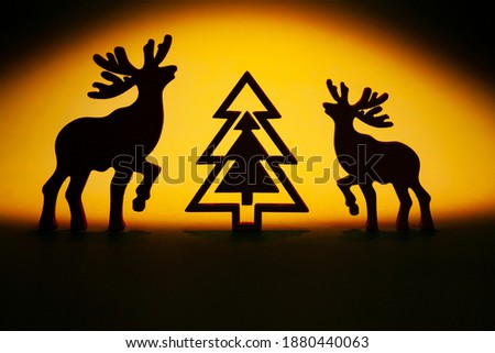 Horizontal conceptual picture with silhouettes of two deers with big horns: adult and young and silhouette of fir on the orange background