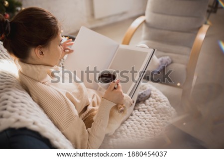 Drinks coffee, reading book near Christmas tree with her pet. Young woman enjoying her domestic life. Home comfort, winter and holidays time concept, warm and cozy atmosphere, lifestyle, New Year.