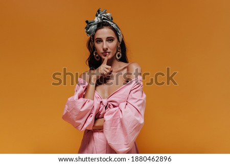 Tanned slim lady in unusual gold earrings, headband and pink modern shirt looking into camera and holding her finger near lips..