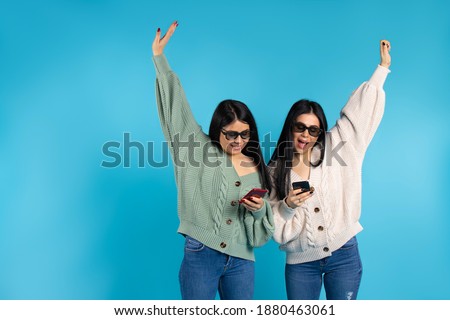 Happy twin girls in 3D glasses with smartphones on a blue background. Hands up and empty side space