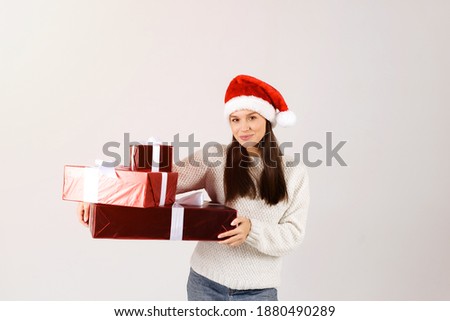 Woman in white sweater holding many gift christmas boxes wearing red santa hat isolated over gray background