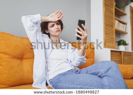 Displeased tired sad woman in casual clothes sit on couch spending time in living room at home. Rest relax good mood leisure concept. Hold using mobile cell phone, making video call put hand on head