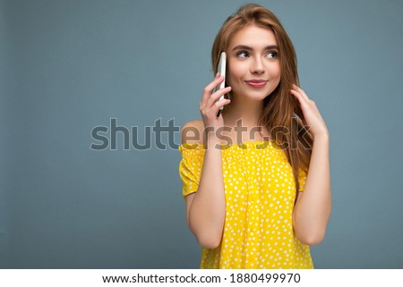 Attractive positive smiling young blonde woman wearing stylish yellow summer dress standing isolated over blue background holding and talking on mobile phone looking to the side
