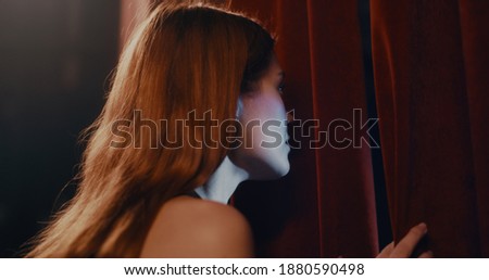Nervous young woman peeking between curtains before performance on stage in theater in evening Royalty-Free Stock Photo #1880590498