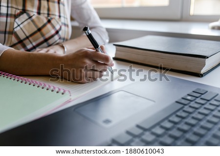 Young woman, student elearning distance training course work at home office and writing content on paper notebook with a pen while studying online course via laptop computer. Close up.