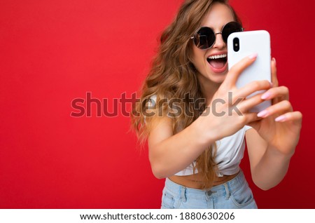 Closeup photo of amazing happy beautiful young blonde woman holding mobile phone taking selfie photo using smartphone camera wearing sunglasses everyday stylish outfit isolated over colorful wall