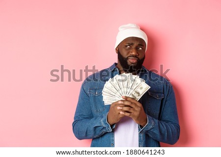 Hesitant african-american man holding money, looking left with doubts and concerns, standing against pink background