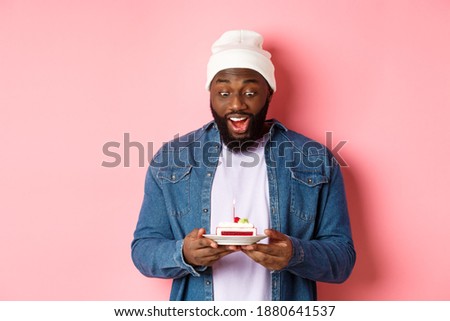 Cheerful african-american guy celebrating birthday, making wish on bday cake with lit candle, smiling happy, standing over pink background