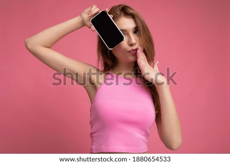 Photo of beautiful positive young blonde woman wearing pink top poising isolated on pink background with empty space holding in hand and showing mobile phone with empty display for cutout looking at