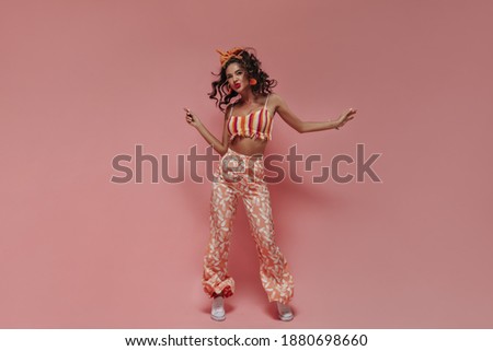 Full length photo of positive wavy haired woman with earrings and bandana in striped cool top, pink modern pants and white sneakers dancing..