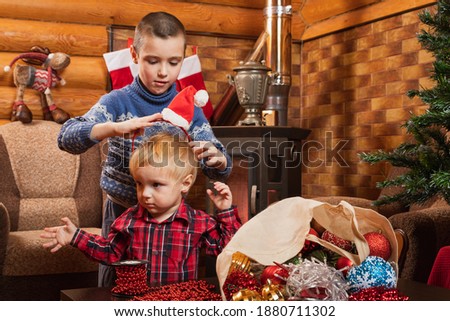 A little boy in a warm knitted sweater puts a Christmas cap on his brother in a country house, in the background there is a decorated Christmas tree, a lighted fireplace, socks for gifts are hung. 