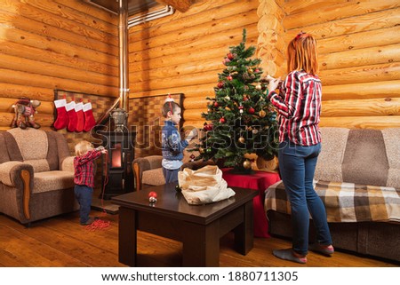 Mom and her sons have a great time preparing for Christmas, smiling in a country house, in the background there is a decorated Christmas tree, a lit fireplace, socks for gifts are hung. 