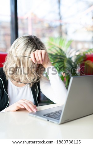 Tired person at the laptop. Woman sits in a cafe and works online, she lowered her head and holds her glasses in hand, vertical photo, selective focus. Overwork, deadline, fatigue, stress, frustration