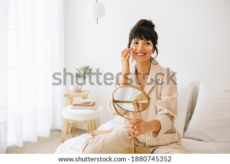 Portrait of woman applying face cream sitting on bed. Smiling young woman in night suit doing skin care at home. Royalty-Free Stock Photo #1880745352