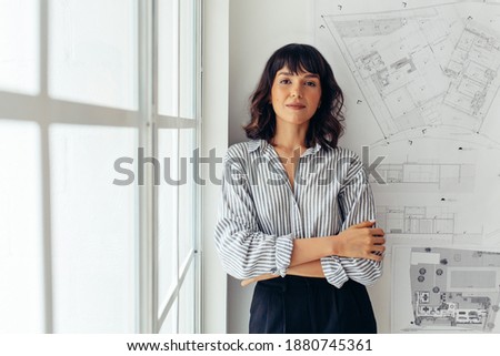Portrait of female architect standing beside architecture drawings in office. Businesswoman standing in office with arms crossed. Royalty-Free Stock Photo #1880745361