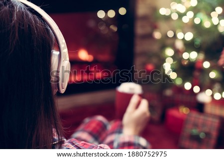 Woman in pajama in headphones with cup of hot cocoa and marshmallow sitting and warming at winter evening near fireplace flame and  christmas tree.
