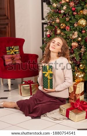 A blonde girl is holding a holiday gift in a light sweater. On the background of the Christmas tree. Festive mood. To give gifts.