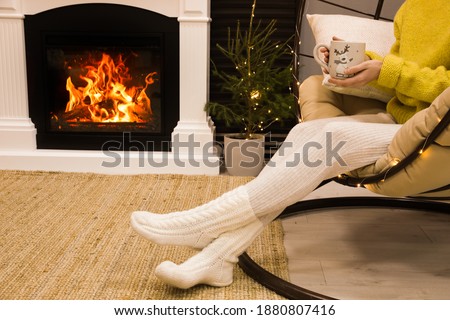 Woman with cup of drink sitting near burning fireplace at home, closeup