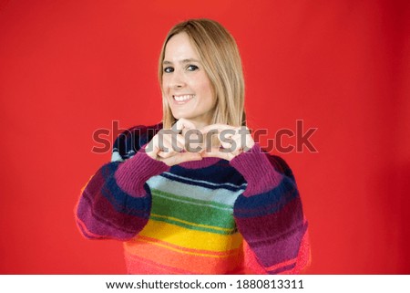 Young woman in colorful sweater making heart figure over red background.