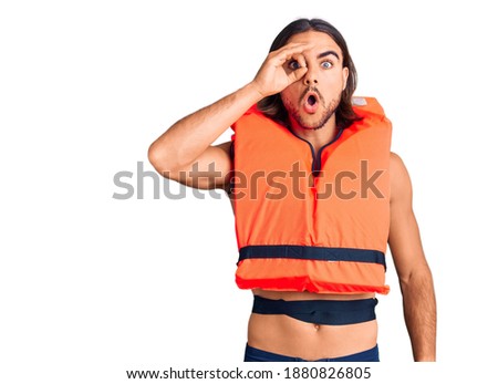 Young handsome man wearing nautical lifejacket doing ok gesture shocked with surprised face, eye looking through fingers. unbelieving expression. 