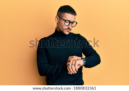 Handsome man with tattoos wearing turtleneck sweater and glasses checking the time on wrist watch, relaxed and confident  Royalty-Free Stock Photo #1880832361