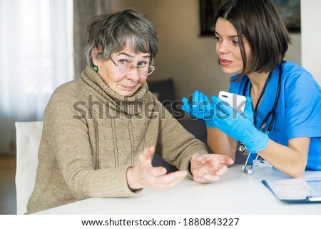 A nurse in a medical suit takes care and explains to an elderly patient how to use applications on a smartphone. Grandmother 80+ years old, does not understand how to communicate with a doctor online.