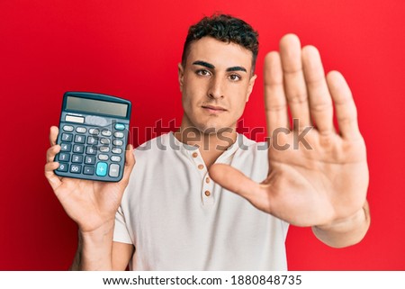 Hispanic young man showing calculator device with open hand doing stop sign with serious and confident expression, defense gesture 