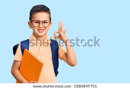 Little cute boy kid wearing school bag and holding book doing ok sign with fingers, smiling friendly gesturing excellent symbol 