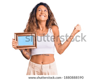 Young hispanic woman with tattoo holding empty frame screaming proud, celebrating victory and success very excited with raised arms 