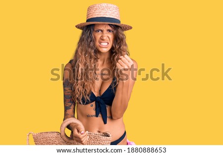 Young hispanic woman with tattoo wearing bikini and hat holding summer wicker handbag annoyed and frustrated shouting with anger, yelling crazy with anger and hand raised 