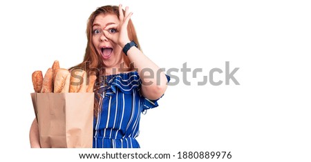Young beautiful redhead woman holding paper bag with bread smiling happy doing ok sign with hand on eye looking through fingers 