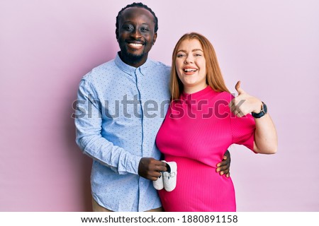Young interracial couple expecting a baby holding shoes smiling happy and positive, thumb up doing excellent and approval sign 