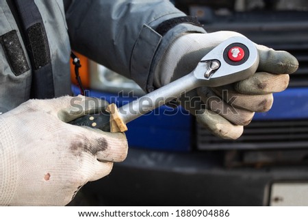 A man wearing work gloves holds a the key for ratchet heads in his hand in the background of a car close-up 