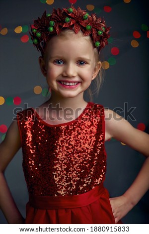 Christmas portrait happy young girl wearing red sequin dress and Christmas wreath. Blond hair and blue eyes posing and fool around in studio on grey background with illumination and boke lights.