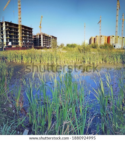 View of a large-scale construction of residential buildings with construction cranes from the opposite side of the lake through the reeds Royalty-Free Stock Photo #1880924995