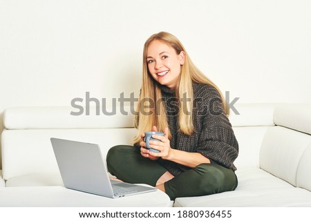Young woman resting on white sofa, wearing warm cozy grey pullover, working with laptop 