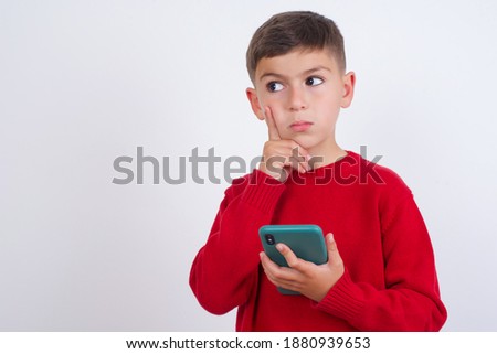 Image of a thinking dreaming Cute Caucasian kid boy wearing red knitted sweater against white wall using mobile phone and holding hand on face. Taking decisions and social media concept.