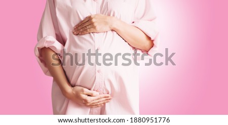 Closeup of beautiful pregnant woman in pink shirt dress holds hands on her belly in white background. Loving mother waiting for baby birth. Pregnancy, maternity, preparation and expectation concept.