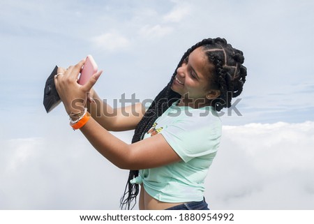 half body shot of one braided hair hispanic beautiful young woman outdoor using her smart phone taking selfie in a cloudy summer day in dominican republic