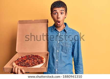 Young african amercian man holding delivery pizza box scared and amazed with open mouth for surprise, disbelief face 