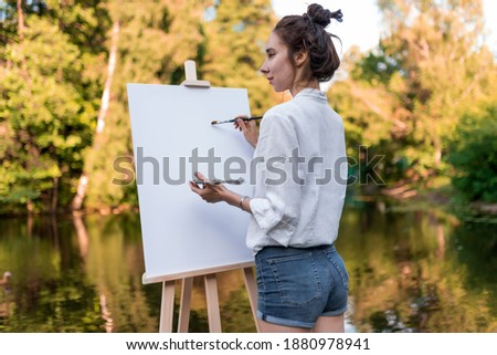 beautiful girl artist, summer park by lake, river pond, draws picture, palette with paints, brush white canvas painting, easel stand. . Green forest background trees water